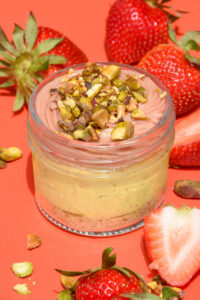 alt="Pistachio Mousse Cheesecake jar with Graham Crust, layered with Strawberry Confit, and topped with Strawberry Ganache and Pistachio Pieces — made from scratch in San Antonio, Texas"