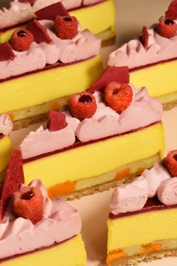 alt="Light Mango Cheesecake layered with Almond Frangipane and sweet Dried Mango Pieces. Topped with tart Raspberry Compote, silky Raspberry Ganache, Fresh Raspberries, and a crisp Raspberry Chocolate Crunch, made from scratch in San Antonio, Texas"