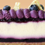 alt="Blueberry cheesecake with graham crust, blueberry jam, vanilla cheesecake filling, blueberry ganache, and white chocolate lime zest crunch made in San Antonio, Texas"