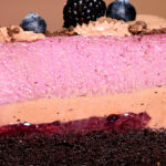 A multi-layered dessert featuring chocolate cake soaked in berry syrup, topped with berry cheesecake, milk chocolate ganache, and mezcal-infused wild berry jam. The dessert is finished with a dusting of chocolate soil and fresh seasonal berries.