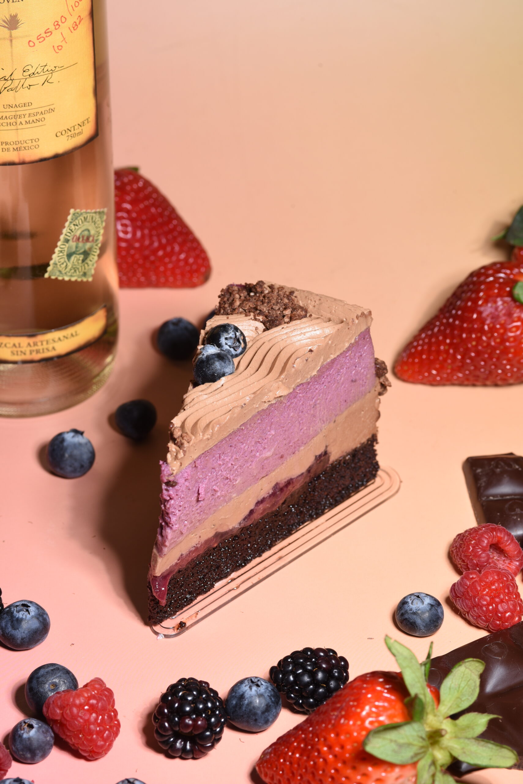 A multi-layered dessert featuring chocolate cake soaked in berry syrup, topped with berry cheesecake, milk chocolate ganache, and mezcal-infused wild berry jam. The dessert is finished with a dusting of chocolate soil and fresh seasonal berries.